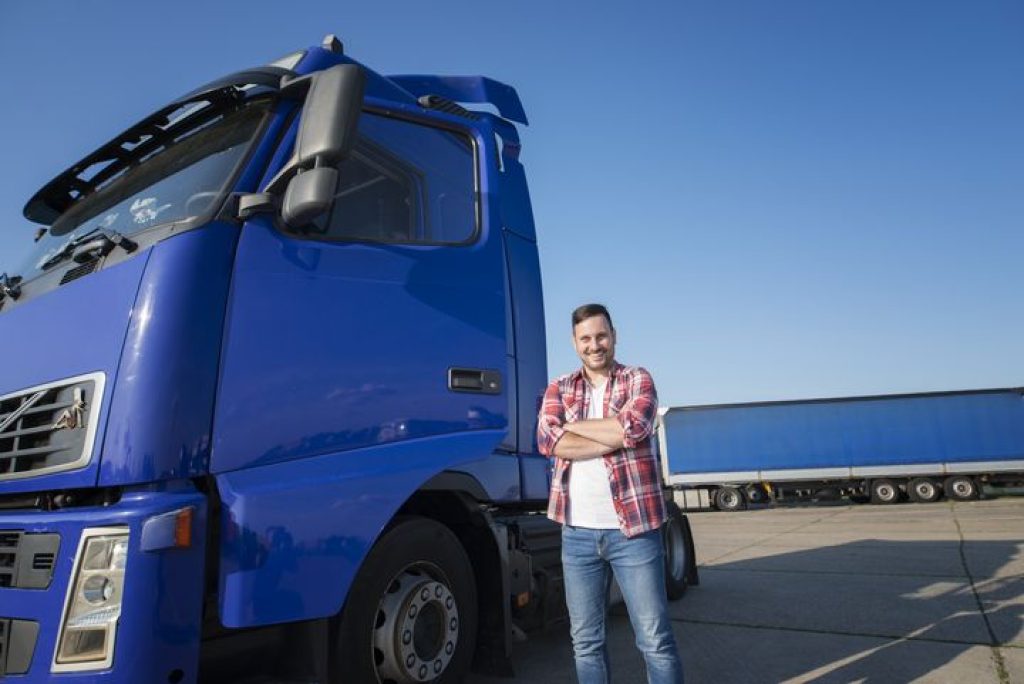 Truck driver in casual clothes standing by his truck with arms crossed at truck stop.